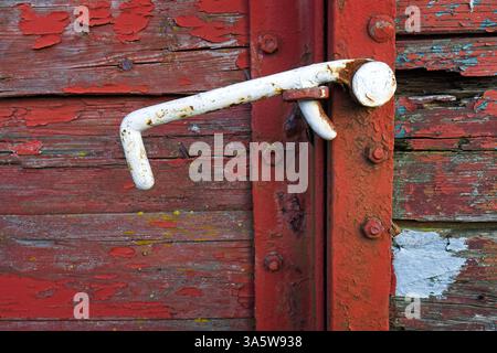 A close-up photograph of a weathered, rusted metal latch on an old wooden door. The peeling red paint on the wooden planks reveals layers of decay. Stock Photo