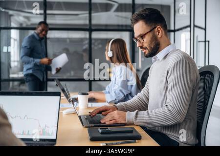 Focused, sitting by laptop. Professional workers are in the modern office together. Stock Photo