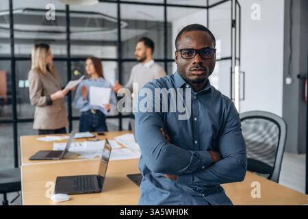 Black man is standing and posing. Professional workers are in the modern office together. Stock Photo
