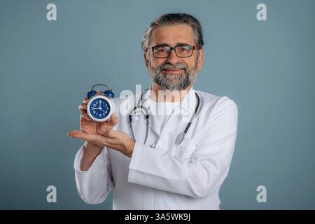 Clock in hands of a doctor. Senior man is standing in the studio against background. Stock Photo