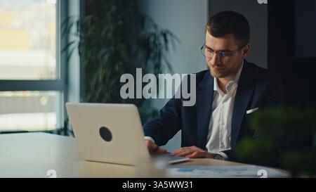 Young businessman, executive, accountant or project manager in a suit working on a laptop at a modern office desk, focused and engaged. productivity Stock Photo