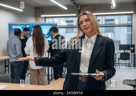 Woman in glasses is standing with notepad. Group of office workers are indoors together. Stock Photo
