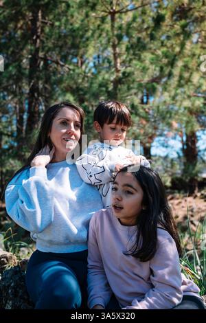 A woman is sitting with two children on a log in a forest. The woman is wearing a gray sweater and the children are wearing pink sweaters. Stock Photo