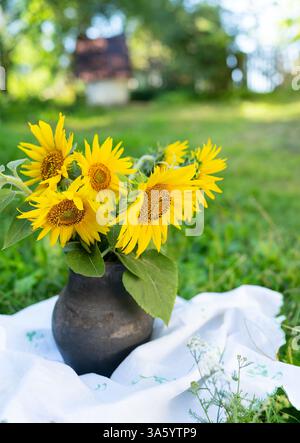 A rustic earthenware vase filled with bright yellow sunflowers sits on a white tablecloth in a sun-drenched garden. Stock Photo