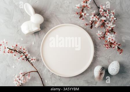 Empty white plate with ceramic bunny, speckled eggs and cherry blossoms on grey background. Easter concept. Top view, flat lay, mockup. Stock Photo