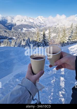 Two friends hands holding paper cups filled with hot chocolate beverage on the top of the a snowy mountain. Winter activity and vacation enjoyment con Stock Photo
