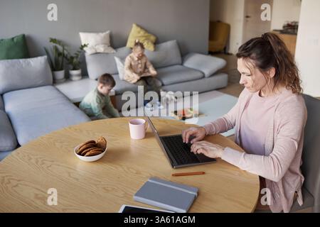 High angle view at single mother using laptop while working from home with two children playing in background, copy space Stock Photo