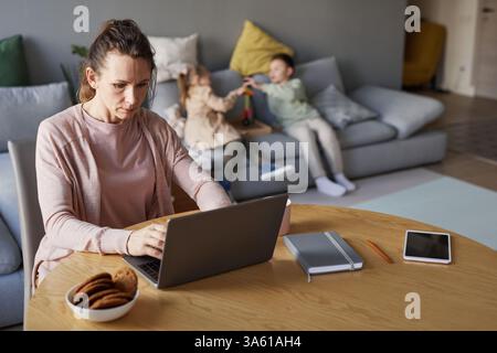 Portrait of single mother using laptop while working from home with two children playing in background, copy space Stock Photo