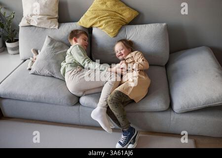 Portrait of cute girl with down syndrome playing with brother and laughing happily while goofing on couch, copy space Stock Photo