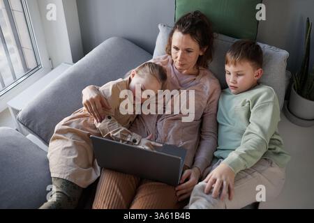 Top down view at caring mother lying on couch embracing son and daughter with down syndrome while watching videos via laptop, copy space Stock Photo