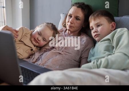 Portrait of caring mother lying on couch embracing son and daughter with down syndrome while watching videos via laptop, copy space Stock Photo
