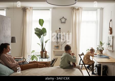 Side view portrait of single mother working from home and using laptop while lying on bed with two children in simple interior Stock Photo