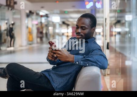 Sitting and holding smartphone. Black man is in the hall of the supermarket. Stock Photo