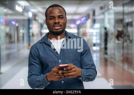 Front view, with smartphone in hand. Black man is in the hall of the supermarket. Stock Photo