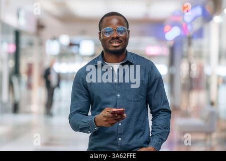 Front view, with smartphone in hand. Black man is in the hall of the supermarket. Stock Photo