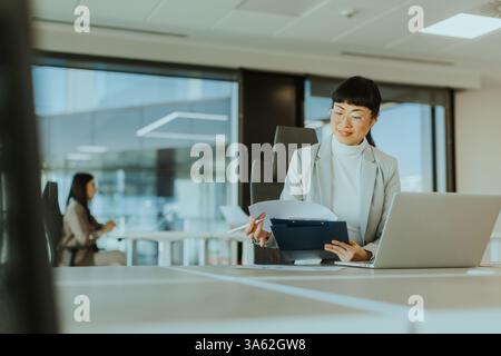 With focused determination, a business professional reviews documents on her laptop in a sleek office setting, while a colleague works quietly in the Stock Photo
