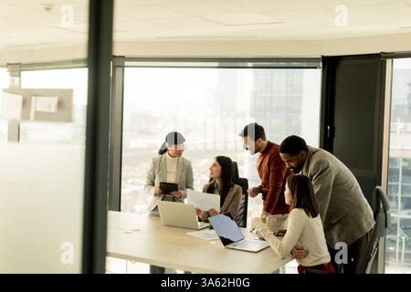 A diverse group of professionals engages in a dynamic brainstorming session around a conference table, sharing ideas while interacting with laptops an Stock Photo