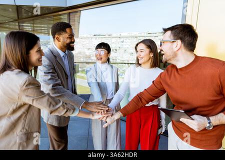 A group of five professionals gathers at a contemporary office space, smiling and joining hands in unity while brainstorming ideas for their upcoming Stock Photo