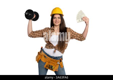 A confident female construction worker wearing a yellow hard hat and tool belt lifts a dumbbell while holding cash, symbolizing financial success, str Stock Photo