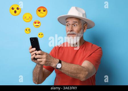 Social media. Surprised senior man using mobile phone on light blue background. Different emojis near gadget Stock Photo