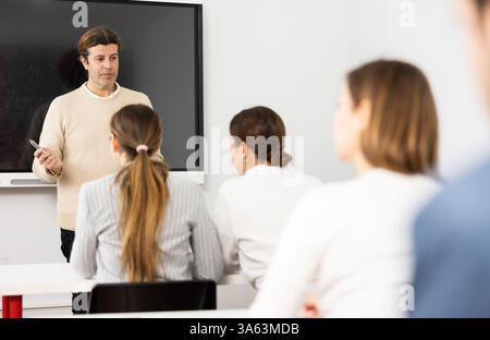 Male business coach explaining topic while standing against group of people sitting at desks in front of him Stock Photo