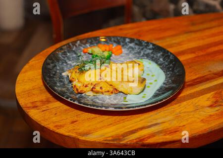 Fried fish fillet with vegetables and sauce on a beautiful plate on a wooden table in a restaurant Stock Photo