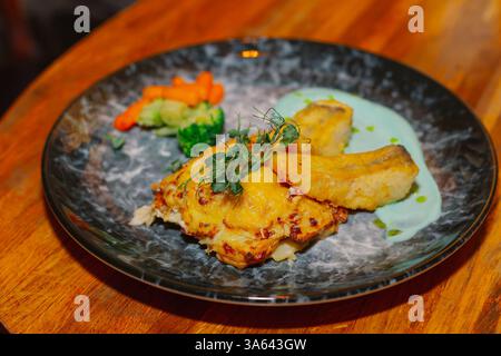 Fried fish fillet with vegetables and sauce on a beautiful plate on a wooden table in a restaurant. dishes from the menu. new cooking recipe Stock Photo