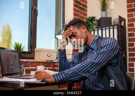 African american businessman leans on table, frustrated while working on marketing strategy presentation on laptop. Black male employee seated at desk with forehead in his hands, overwhelmed by work. Stock Photo