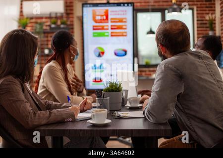 Coworkers seated at table with notepads, taking notes and discussing business project analytics shown on digital monitor. Professional team analyzing financial charts on led screen in startup office Stock Photo