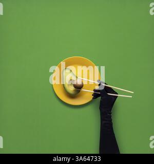 Creative concept photo of avocado with hand and chopsticks on painted plate on green background Stock Photo