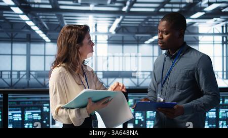 Cheerful admins meeting in data center to review reports and files, doing handshake. Server room employee saluting colleague, doing troubleshooting to prevent downtime, camera B Stock Photo