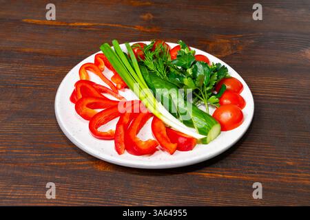 A variety of fresh vegetables are artistically arranged on a white plate. Red bell peppers, cherry tomatoes, green onions, cucumber, and cilantro crea Stock Photo