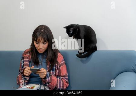 Young woman using smartphone while eating on blue sofa with black cat sitting next to her Stock Photo