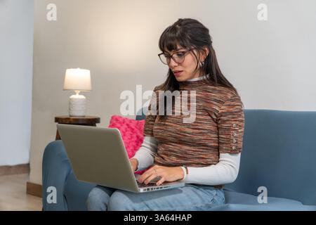 Focused latin woman working remotely from home office using laptop on sofa Stock Photo
