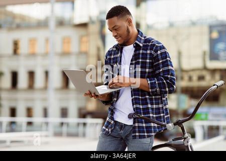 Black man using laptop, standing in downtown with bike Stock Photo