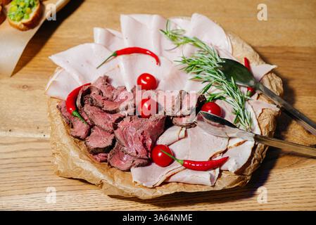 Top view of a charcuterie platter featuring sliced roast beef, ham, cherry tomatoes, chili peppers, and rosemary garnish. Stock Photo
