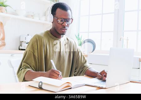 Young black man wearing headphones and glasses, taking notes in notebook while using laptop for online education, working remotely or attending online meeting or webinar in bright kitchen Stock Photo