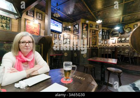A woman sitting thoughtfully in a cozy pub interior with wooden decor, wearing a pink scarf. Two beer glasses are on the table, creating a warm and ca Stock Photo