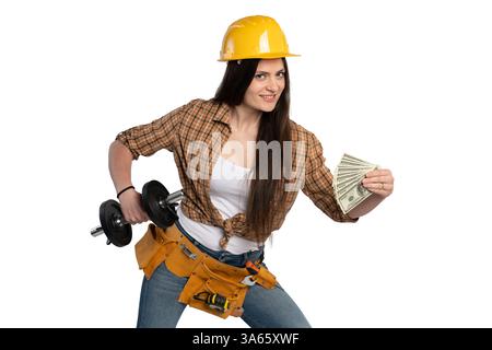 A smiling female construction worker wearing a yellow hard hat and tool belt holds a dumbbell in one hand and cash in the other, symbolizing financial Stock Photo