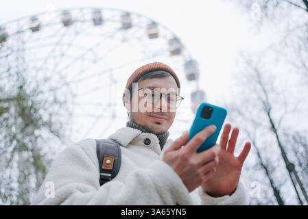 Young man wearing glasses and winter clothes using mobile phone app near a ferris wheel, enjoying his vacation in an amusement park during winter Stock Photo
