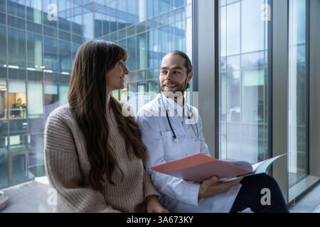 Male doctor in hospital clinic wearing medical uniform reviewing papers with patient Stock Photo