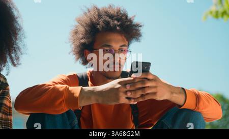 Two African American students friends woman girl female man male guy together gadget addict using mobile phones scrolling smartphones social media sit Stock Photo