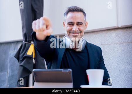 Smiling man pointing at camera while working with tablet and coffee in outdoor cafe Stock Photo