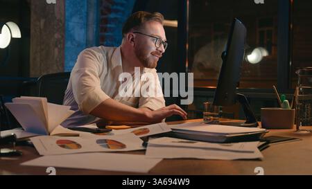 Caucasian man business guy businessman male workaholic in dark night office late time working computer celebrate smiling satisfied finish online work Stock Photo