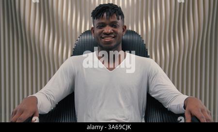 African American ethnic happy smiling positive cyber man in white room futurism studio indoors male guy looking at camera smile carefree sitting in Stock Photo