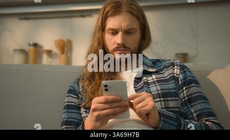 Calm serious Caucasian man guy male user gadget addict indoors at home couch comfortable sofa using mobile phone scrolling social apps media reading Stock Photo