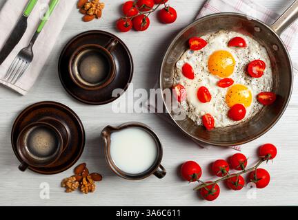Two fried eggs with cherry tomatoes in a pan, two cups of coffee, milk, nuts and cherry tomatoes branches are composing a delicious breakfast on a white wooden table Stock Photo