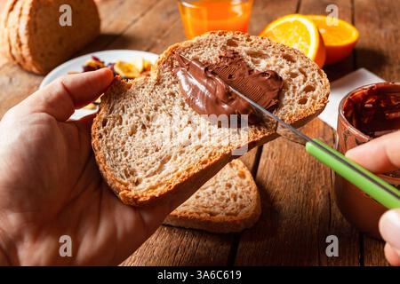 Close up of hands spreading chocolate hazelnut cream on whole wheat toast, with orange juice and sliced oranges in the background, creating a healthy breakfast scene Stock Photo