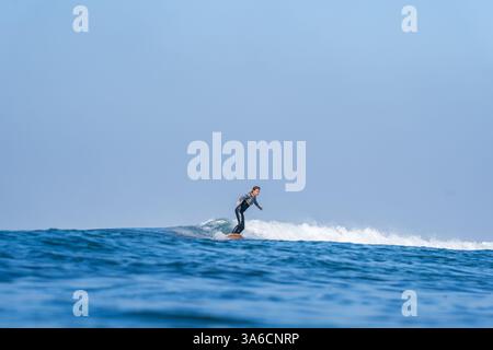 A young woman in a wetsuit rides a surfboard on a small wave.  Blue clear sky, blue sea. Stock Photo