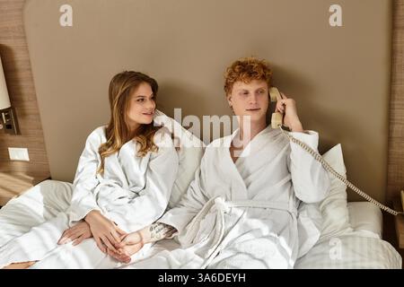 A young couple in hotel robes smiles together on a bed while one talks on the phone. Stock Photo
